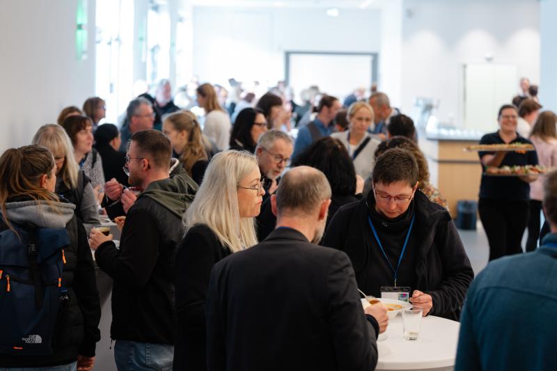 Viele Menschen stehen in einem hellen Raum zusammen, reden und essen an Stehtischen.