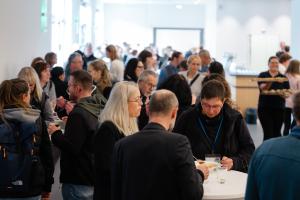 Viele Menschen stehen in einem hellen Raum zusammen, reden und essen an Stehtischen.